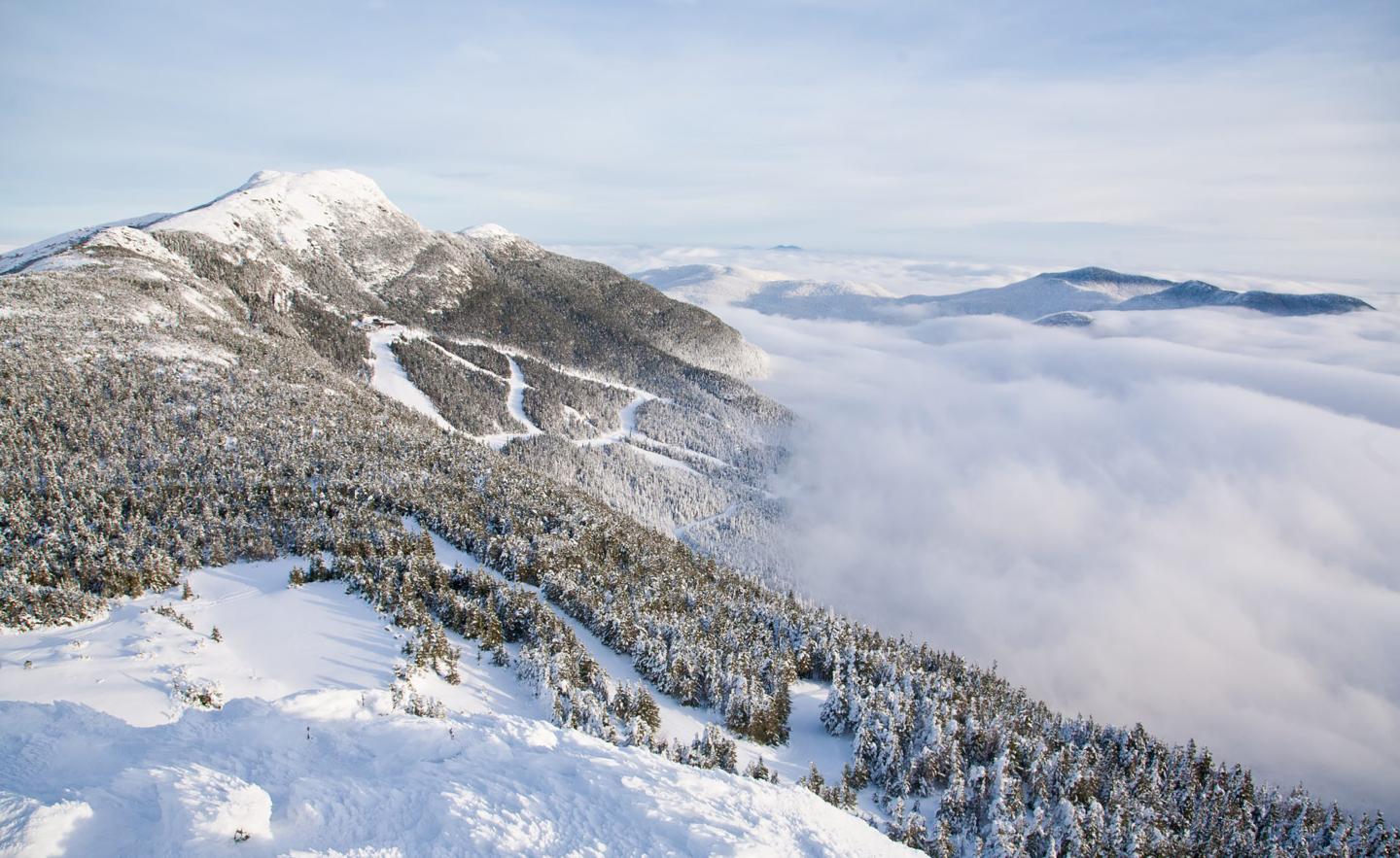 Snowy mountain landscape with clouds below and ski trails visible.