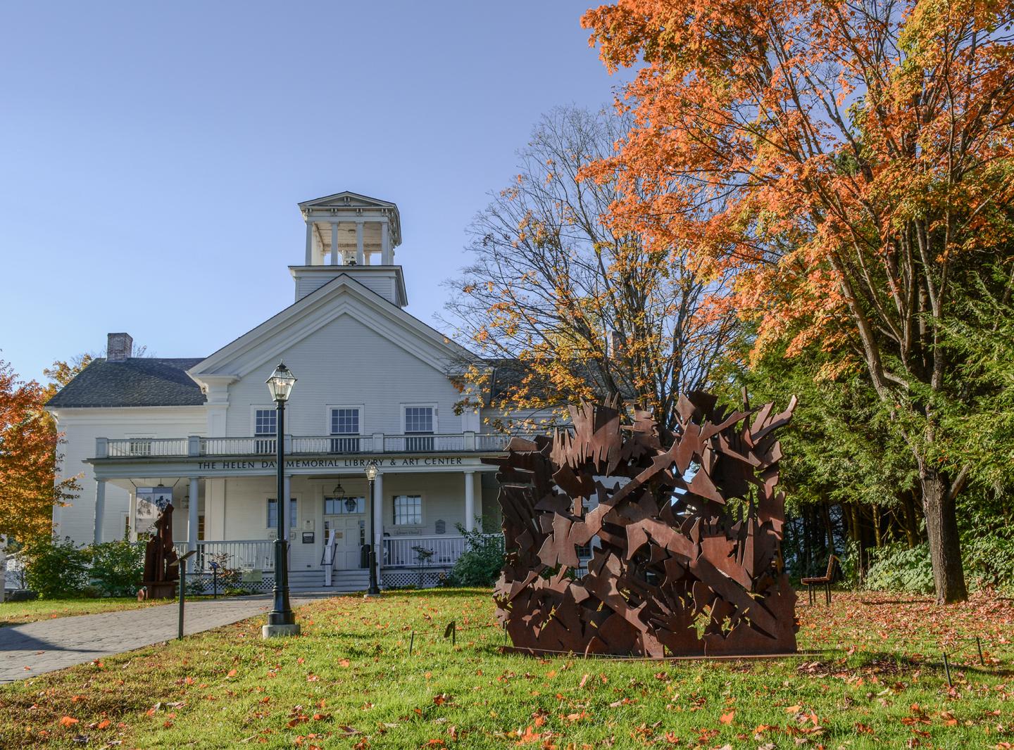 White building with a tower, surrounded by autumn trees and metal sculpture in the foreground.