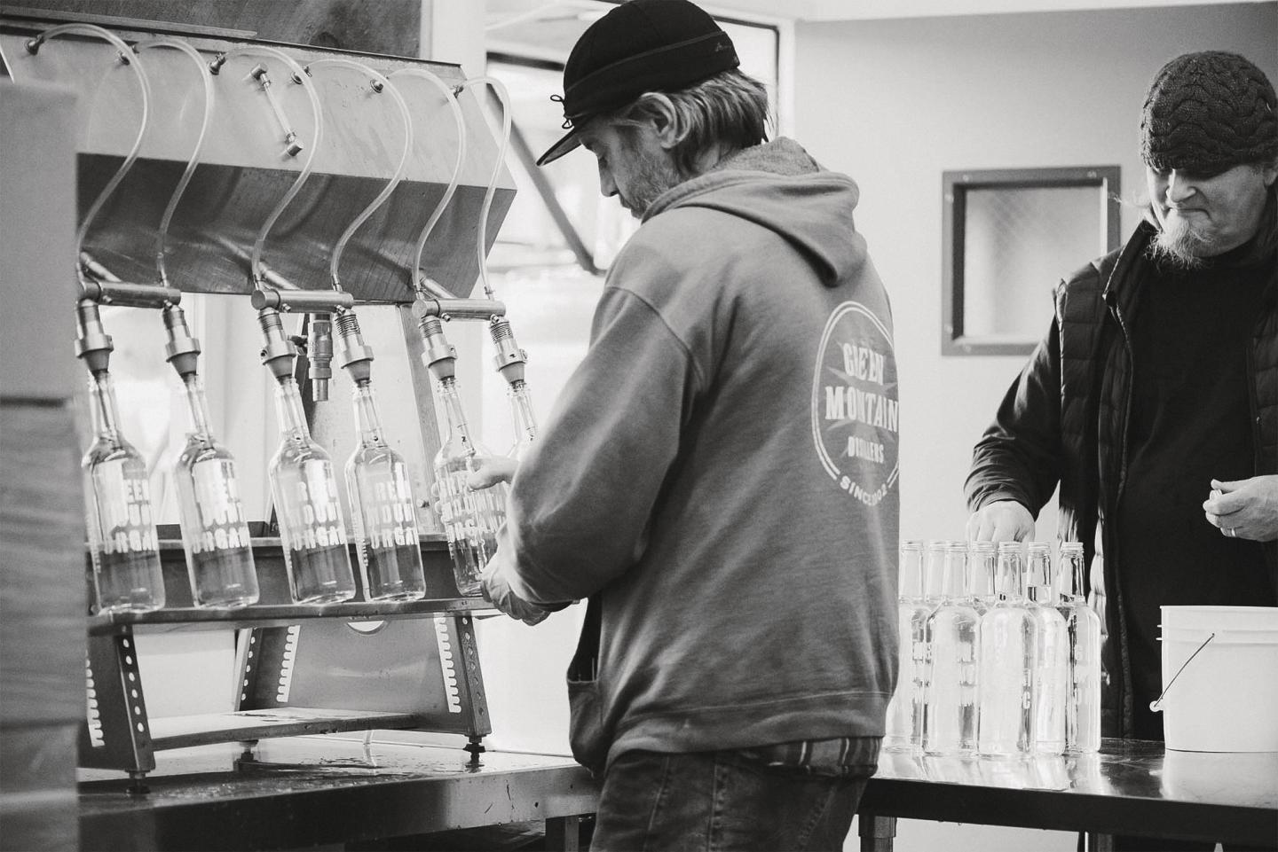 Men bottling liquid in a factory setting, wearing casual clothes and hats.