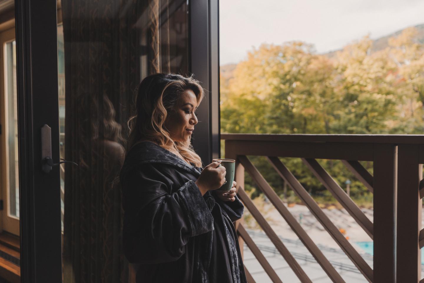 Woman holding a cup on a balcony, surrounded by autumn trees.
