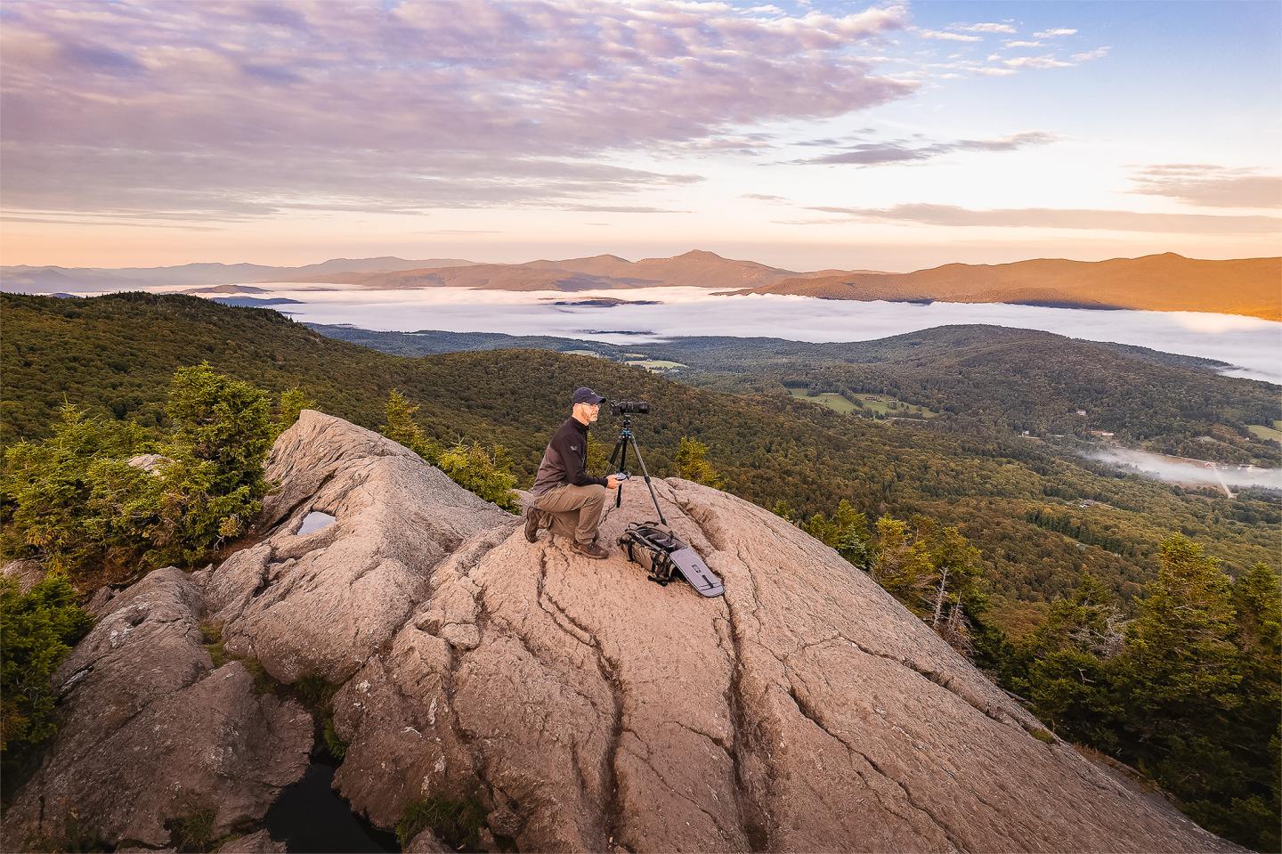 Photographer on rocky peak with camera, sunrise landscape in background.