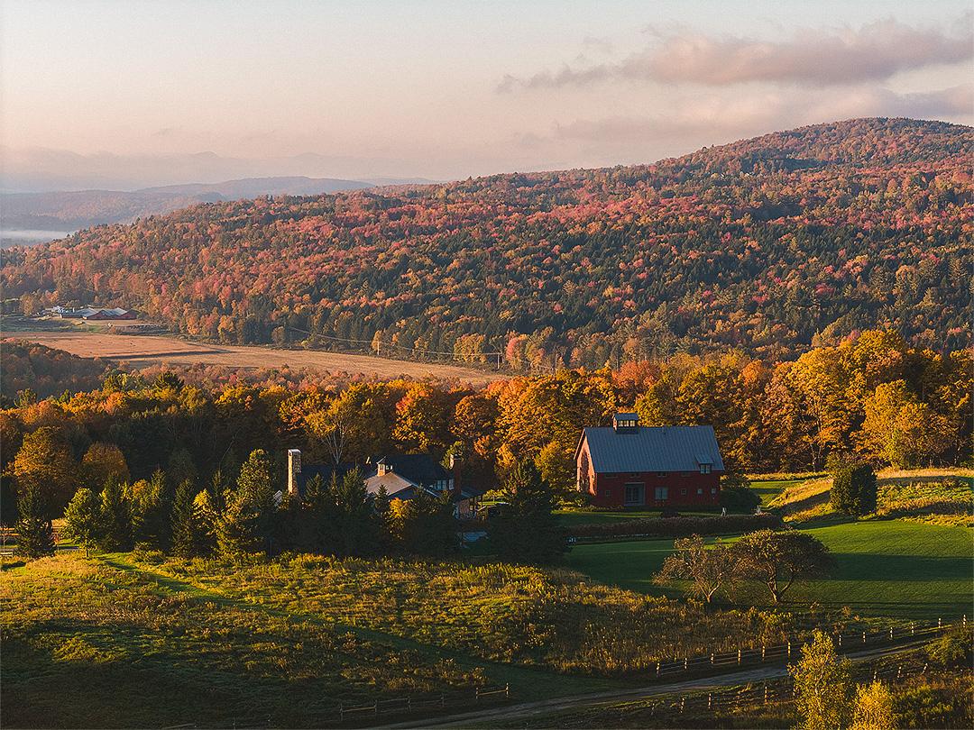 Mountain landscape with a red barn, surrounded by autumn trees at sunset.