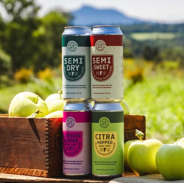 Four colorful beverage cans stacked outdoors with green apples and mountains in the background.