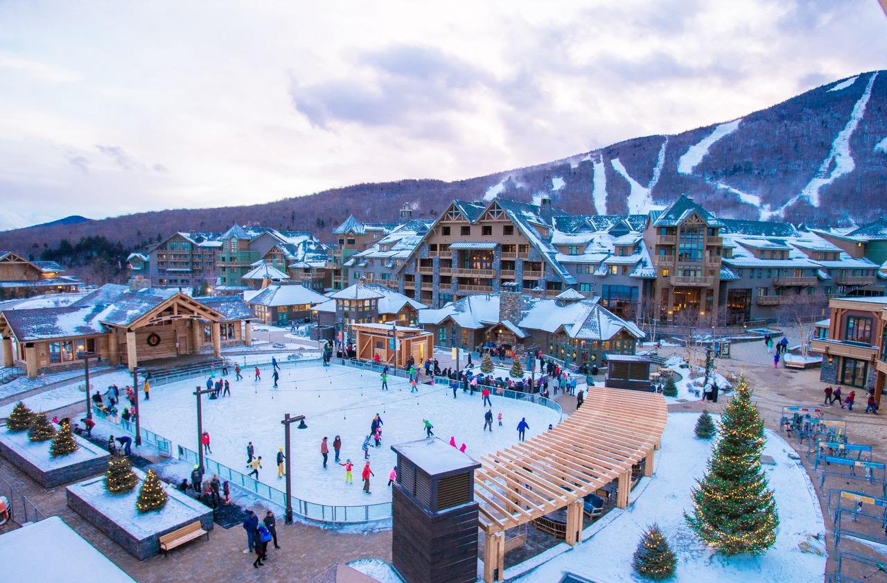 Snowy resort with ice rink, people skating, and mountains in the background.