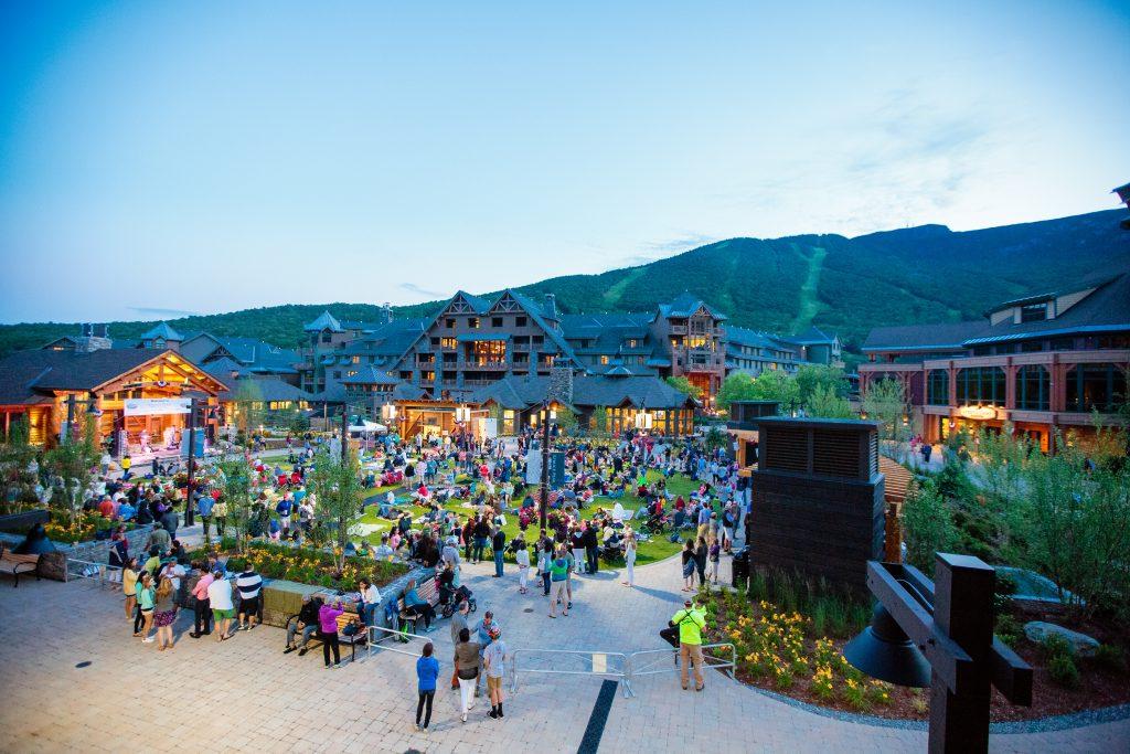 Crowd gathers in a lively plaza with mountain backdrop at dusk.