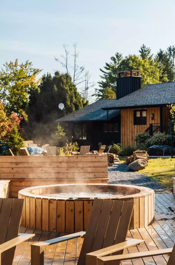 Wooden outdoor hot tub with steam, surrounded by chairs and greenery. Rustic cabin in the background.