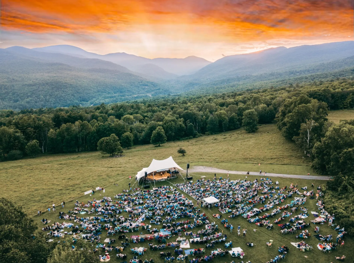 Outdoor concert at sunset, large crowd on grassy field, mountains in background.